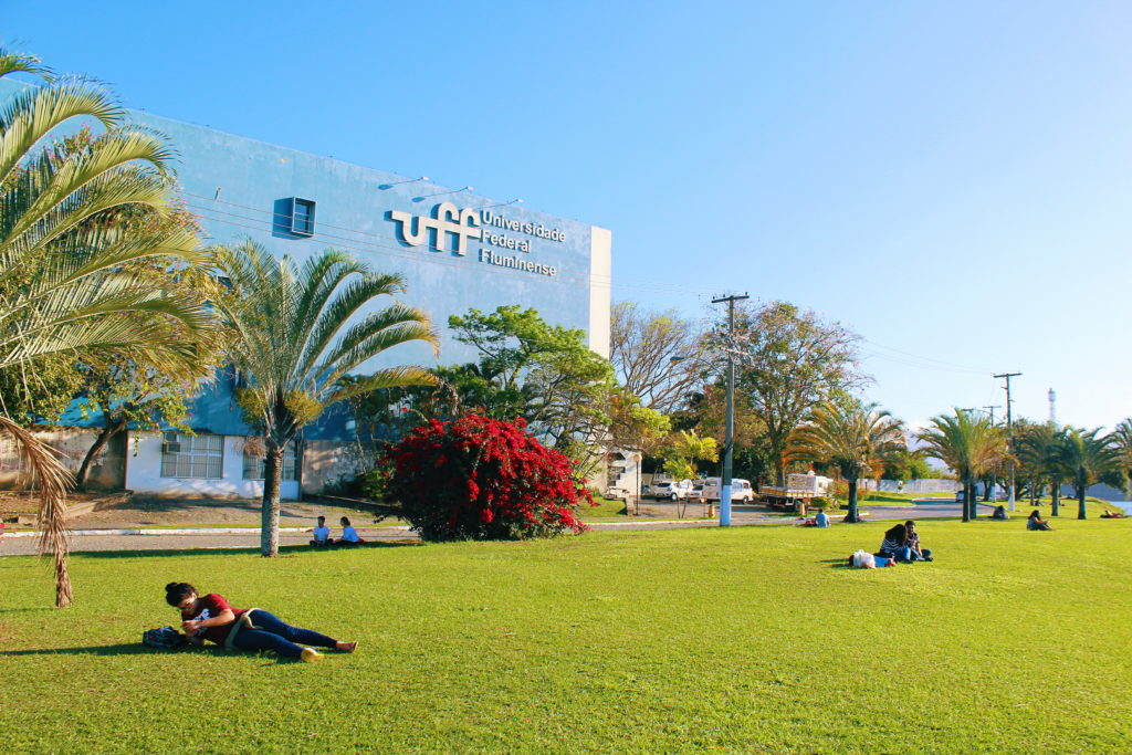 Uma foto da orla da uff com pessoas na grama, árvores e um prédio da UFF atrás.